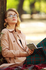 Young lady in retro style, student in park in nature wearing glasses and holding notepad in her hands