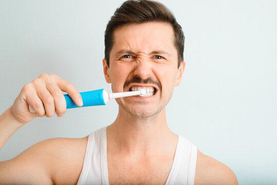 Portrait of handsome man persistently, strongly brushing his teeth with modern electric toothbrush and looking at camera indoors. Young man daily oral hygiene and teeth whitening