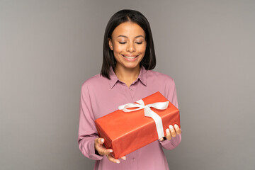 Surprised african american woman holding gift box for christmas or new year celebration. Happy young female looking at present for valentines day, birthday or 8 march wrapped in decorative red paper