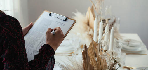 Unrecognizable Woman Hosting Christmas Dinner Party at Home.
Close up photo of female hands writing notes on a clipboard while standing in a restaurant and looking at weeding or birthday table set up.