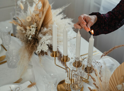 An Anonymous Woman Setting Up Christmas Dinner Table At Home. 

Close Up Photo Of Female Hands Lights The Candle On Golden Candlestick Holder And Arranging Table For Birthday Party.