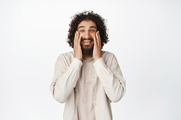 Image of smiling surprised man with curly hair, looking amazed and touching his face, standing in...
