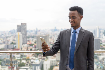Portrait of handsome African businessman wearing suit and tie at rooftop in city while using mobile phone