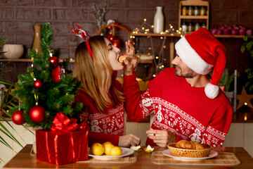 happy lovers a man and a girl in red sweaters in the kitchen with a Christmas tree feed each other tangerines at home and celebrate New Year or Christmas rejoicing and smiling