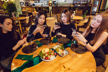 A group of women friends are sitting at a table in a restaurant and using smartphones.