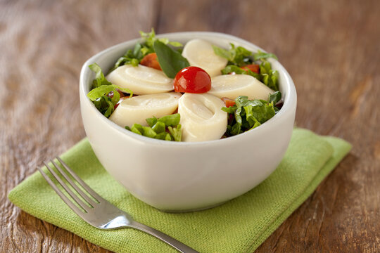 Salada de alface picada com tomate cereja e palmitos fatiados em cumbuca no fundo de mesa r&uacute;stica.