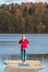 Beautiful blonde hair girl doing yoga on a wooden pier on the lake in autumn with forest on background, vertical