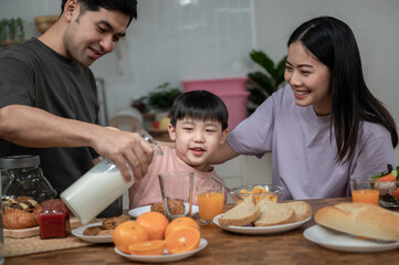 Happy Asian family enjoying breakfast together on dinning table. Young Asian father pouring a glass of milk to his son while having breakfast in the kitchen. Happy Asian family lifestyle concept.