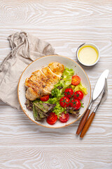 Healthy green vegetable salad with grilled chicken breast fillet on ceramic plate with olive oil on the side on white wooden kitchen table top view flat lay, diet food and clean eating concept
