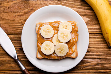 Toast with peanut butter and banana on white ceramic plate with knife on rustic wooden background,  jar with spoon, top view flat lay - Powered by Adobe