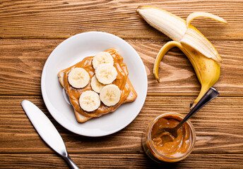 Toast with peanut butter and banana on white ceramic plate with knife on rustic wooden background,  jar with spoon, top view flat lay