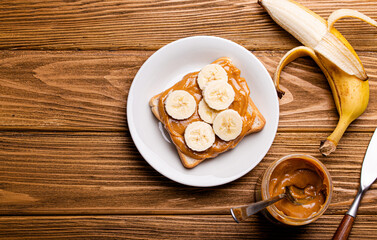 Toast with peanut butter and banana on white ceramic plate with knife on rustic wooden background, top view flat lay, space for text