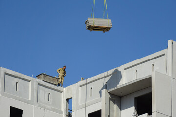 Construction of a multi-storey building. An unsecured builder stands on the edge and looks down. The plates are lifted up by a crane.