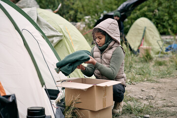 Little girl in warm jacket unpacking box with new clothes while sitting by refugee tent