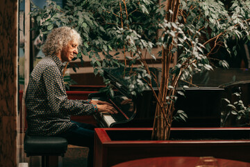  Portrait of gray-haired senior man with curly long hair is sitting at piano and playing music, enjoying pleasant evening at hotel