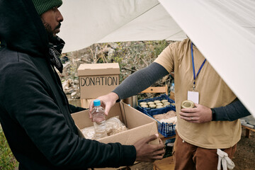 Young refugee man holding box while volunteer putting there food and water for the family of...