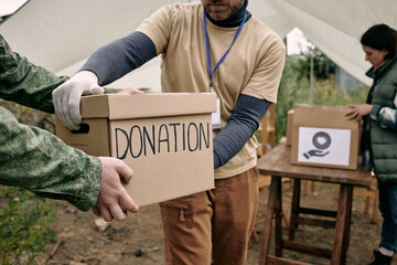 Young man in gloves taking package with donation against female unpacking other boxes