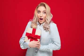 Surprised girl holding gift box - present with ribbon, isolated on red background. Young woman with...