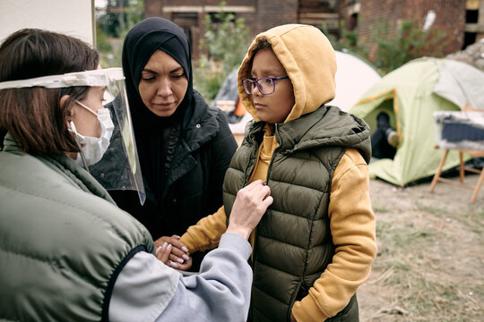 Young volunteer in protective mask and screen examining little girl in refugee camp