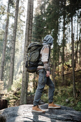 Back view of male hiker backpacker in highlands mountains. Man with heavy backpack relaxing standing on the rock looking around forward on pine trees.