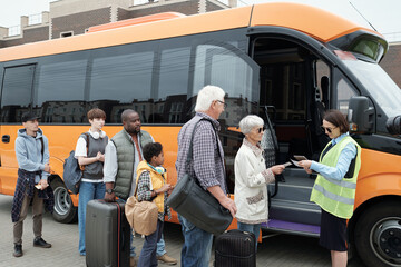Young bus conductor checking tickets by open door of vehicle with line if passengers in front