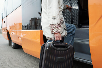 Contemporary senior passenger with suitcase putting leg on staircase while entering bus