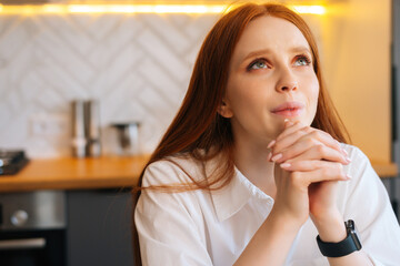 Close-up face of sad young woman praying holding folded hands in front of face at home, looking up. Depressed female put hands in prayer praying excitedly with hope, thinking about something.
