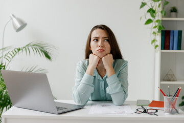 Young bored disappointed frustrated sad employee business woman 20s in blue shirt prop up chin sit...