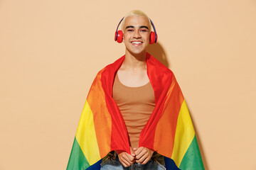 Young satisfied blond latin gay man with make up in beige tank shirt headphones wrapped in rainbow...