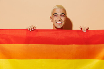 Young smiling minded blond latin gay man with make up in beige tank shirt hiding with rainbow flag...