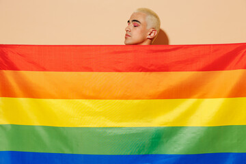 Side view young smiling bisexual blond latin gay man with make up in beige tank shirt hiding with...