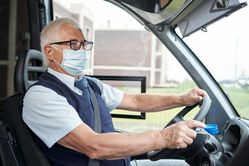 Senior bus driver in casualwear, eyeglasses and protective mask steering and looking through front...
