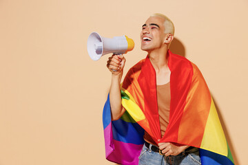 Young blond latin gay man with make up in beige tank shirt wrapped in rainbow flag hold shout aside...