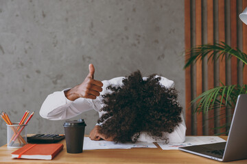 Young tired african american employee business man wearng classic shirt put head down on table sleeping show thumb up sit work at white office desk with pc laptop inside Achievement career concept