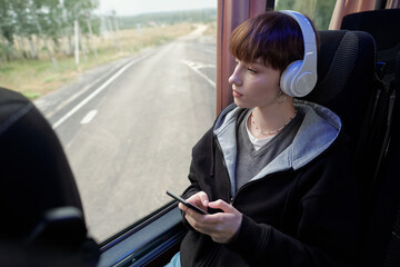 Young serene woman in headphones looking through window of bus while traveling to another city