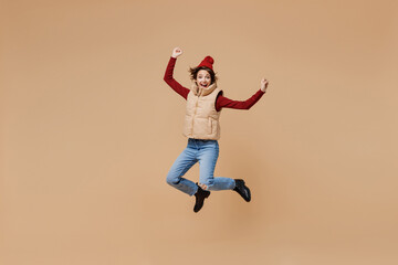 Fototapeta na wymiar Full body overjoyed happy young woman 20s wears red turtleneck vest beret jump high do winner gesture clench fist isolated on plain pastel beige background studio portrait. People lifestyle concept.