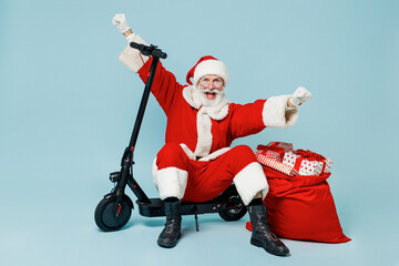 Full body tired old Santa Claus man in Christmas hat red suit clothes sit scooter with gifts bag do winner gesture isolated on plain blue background studio. Happy New Year 2022 merry ho x-mas concept.