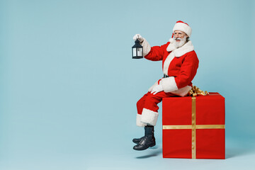 Full size old Santa Claus man in Christmas hat red suit clothes sitting on big gift present box hold lantern isolated on plain blue background studio Happy New Year 2022 merry ho x-mas holiday concept