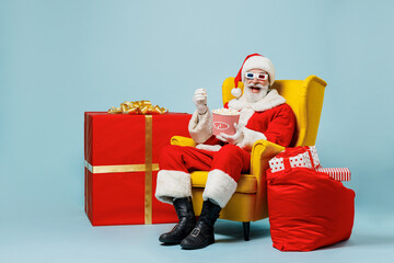 Full body old Santa Claus man 50s in Christmas hat red suit 3d glasses sit in chair eat popcorn watch movie isolated on plain blue background studio Happy New Year 2022 merry ho x-mas holiday concept