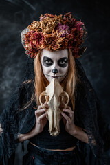 halloween red-haired Caucasian woman in makeup, a witch costume, with a sewn-on mouth, horns, dried flowers on her head, in dark smoky room holds wolf skull in front of her, looks at camera, conjures