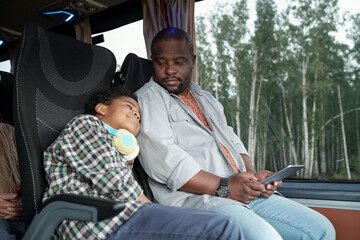 Young African man with digital tablet looking at his cute sleeping son during bus ride