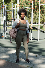 Young happy African sportswoman with handbag standing on sportsground before training