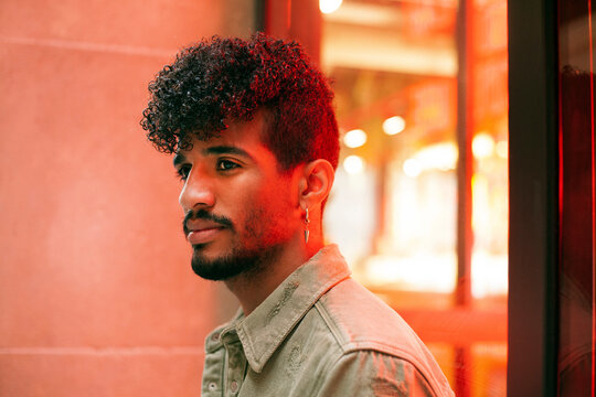 Portrait of young biracial curly haired man with orange neon light reflection on face. Looking to the side. Millennial with beard and earring. Urban style. Horizontal image.