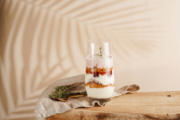 Tall glass with a layered creamy yoghurt dessert trifle with cookies, amaretti, cherries and thyme on a linen cloth on a rustic wooden table at a tropical setting