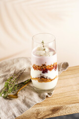 Tall glass with a layered creamy yoghurt dessert trifle with cookies, amaretti, cherries and thyme on a linen cloth on a rustic wooden table at a tropical setting