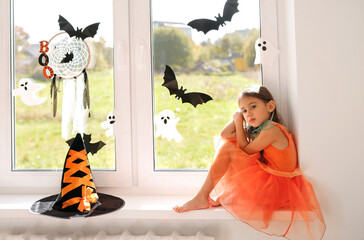 A girl is sitting on the windowsill waiting for Halloween. Holiday party decorations: bats, hat...