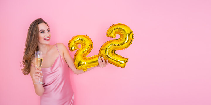 Young happy girl holds gold foil balloon and glass of champagne on pink background BIRTHDAY PARTY