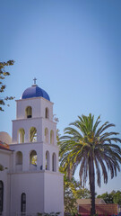 Fototapeta na wymiar Small stucco Catholic church with bell tower surrounded by palm trees in sunny beach town on the Pacific Ocean coast