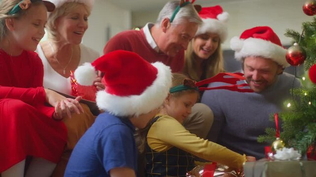 Girl unwrapping present of soft toy dog as multi-generation family exchange gifts around christmas tree at home - shot in slow motion