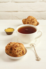 Oatmeal cookies with walnuts and raisins on small saucers and a cup of black tea, standing on a white wooden table against a white brick wall. Closeup
